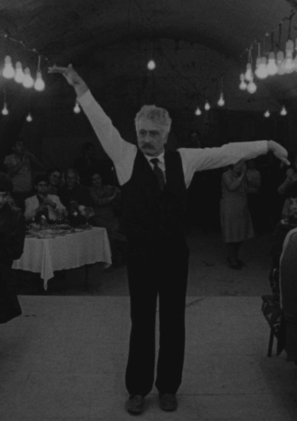 Ein Mann mit Weste und Krawatte steht in einem schwach beleuchteten Raum, einen Arm erhoben und den anderen zur Seite gestreckt, als würde er tanzen. Die Leute sitzen an blumengeschmückten Tischen und schauen zu, und hängende Glühbirnen erhellen die Szene.