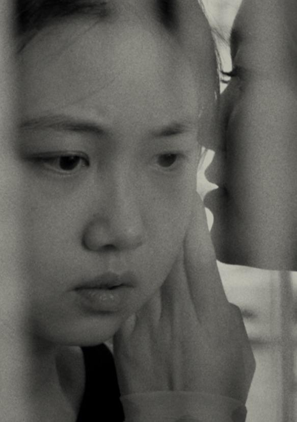 Eine Schwarz-Weiß-Nahaufnahme einer jungen Person, die nachdenklich dreinschaut, während eine andere Person ihr ins Ohr flüstert und dabei sanft ihr Gesicht berührt. Vertikale Unschärfen oder Linien verdecken teilweise die Szene.